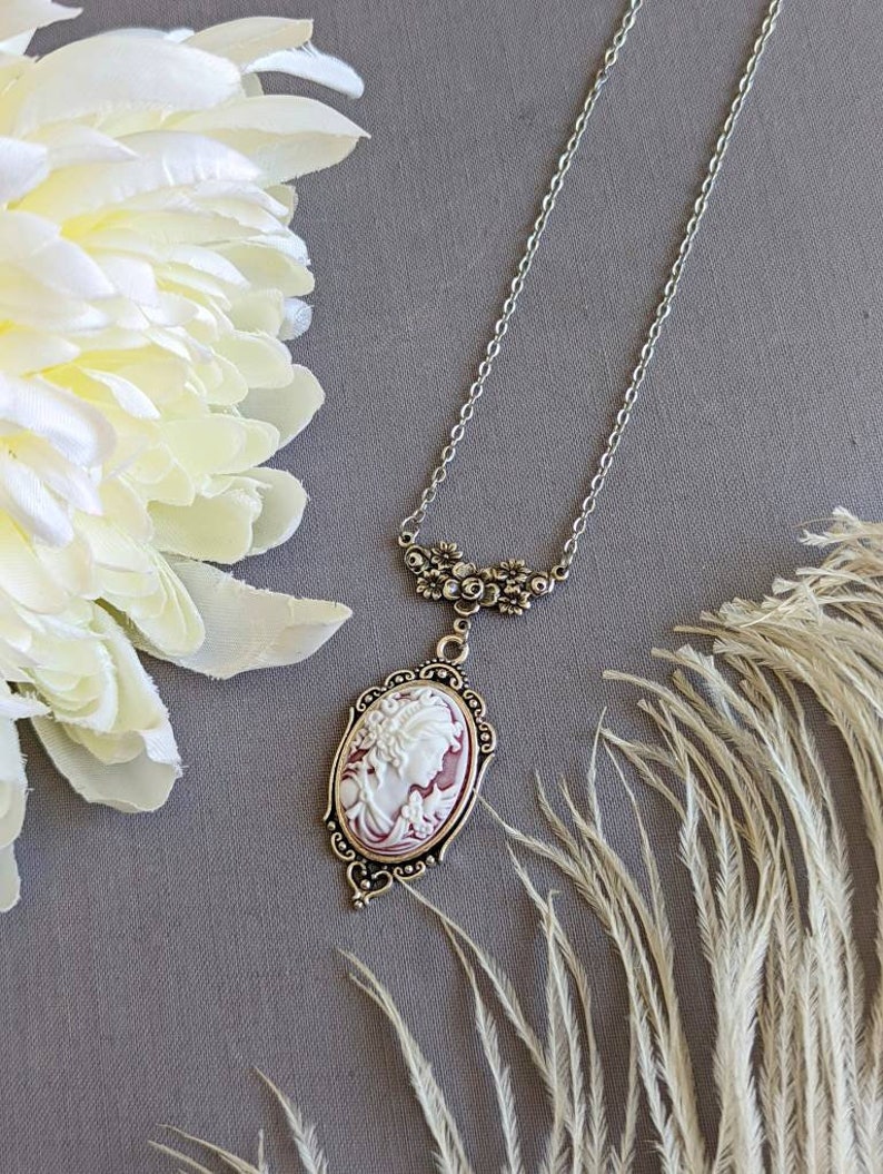 Ornate silver Cameo necklace with white and wine cameo displayed on grey background with flowers and feathers surrounding it.