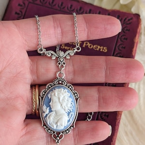 A close up of a blue and white cameo necklace with an ornate silver pendant and tulip connector being held in a woman's hand.