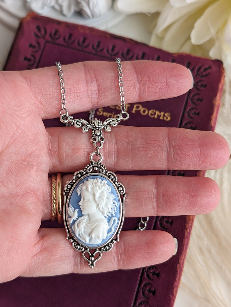 A close up of a blue and white cameo necklace with an ornate silver pendant and tulip connector being held in a woman's hand.