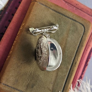 Open floral embossed oval shiny silver locket with bow style pin displayed on a stack of vintage books.