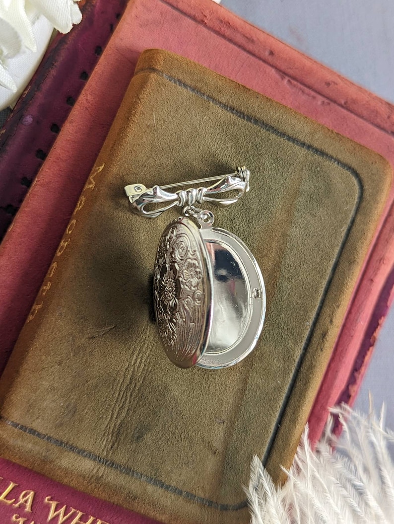 Open floral embossed oval shiny silver locket with bow style pin displayed on a stack of vintage books.