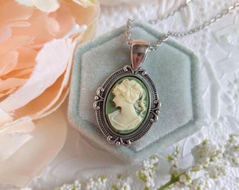 May include: An oval pendant necklace featuring a cream-colored cameo profile of a person set against a sage green background. The cameo is encased in an ornate, silver-toned metal frame with decorative scrollwork and hangs from a silver chain.