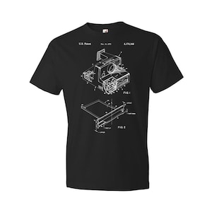 May include: Black t-shirt with a white graphic of a Polaroid camera patent design. The patent number is 4,174,164. The text "U.S. Patent" and "FIG. 1" and "FIG. 2" are also included in the design.