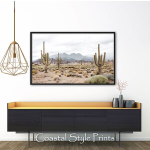 May include: A framed photograph of a desert landscape with tall cacti and mountains in the background. The photo is in a black frame and is hanging on a white wall above a black dresser with a light wood top.