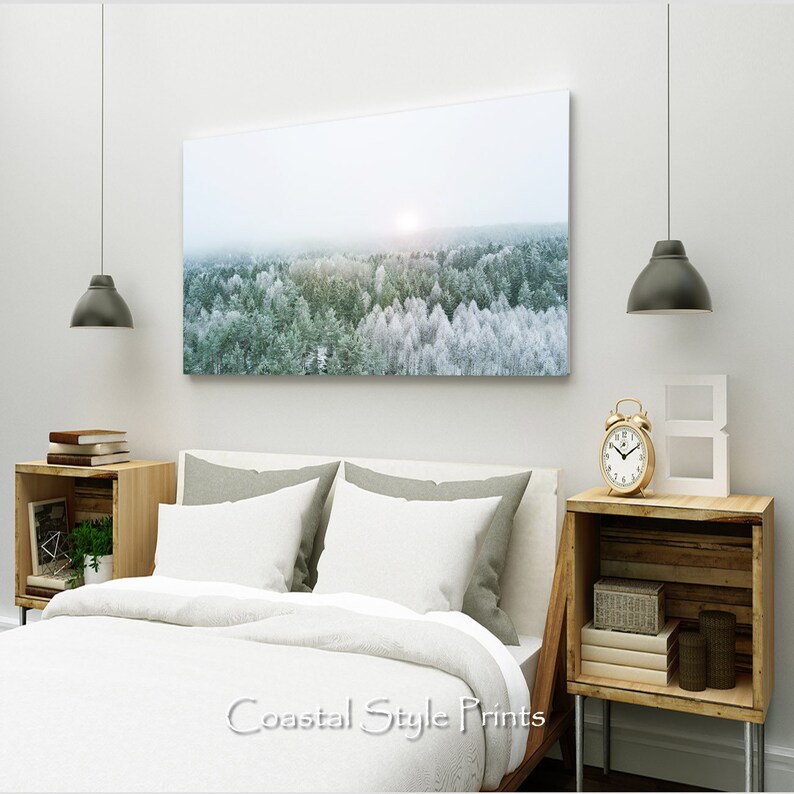 May include: A white bed with a white duvet cover and two gray pillows. A framed print of a snowy forest hangs above the bed. A wooden nightstand with a gold alarm clock and a white letter 'B' sits next to the bed.