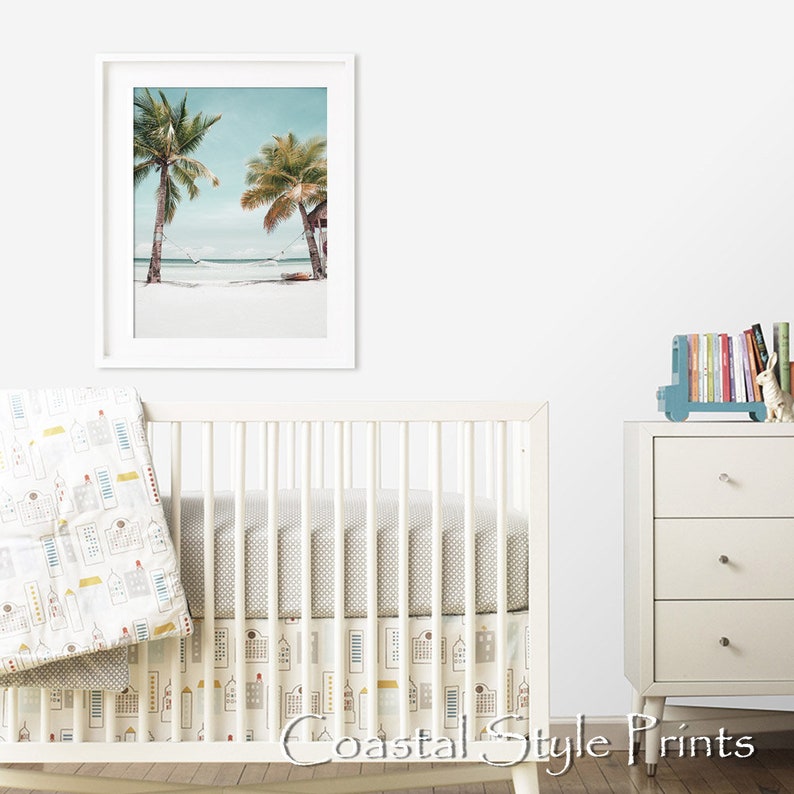 Op de afbeelding: Een wit babybedje met een grijs-wit gestreept lakentje en een wit-gele gestreepte rok. Een ingelijst printje van twee palmbomen op een strand hangt aan de muur achter het babybedje. Een witte commode met drie lades staat rechts in de afbeelding. Een blauwe speelgoedtruck met boeken erop staat op de commode. De tekst "Coastal Style Prints" staat onderaan de afbeelding.