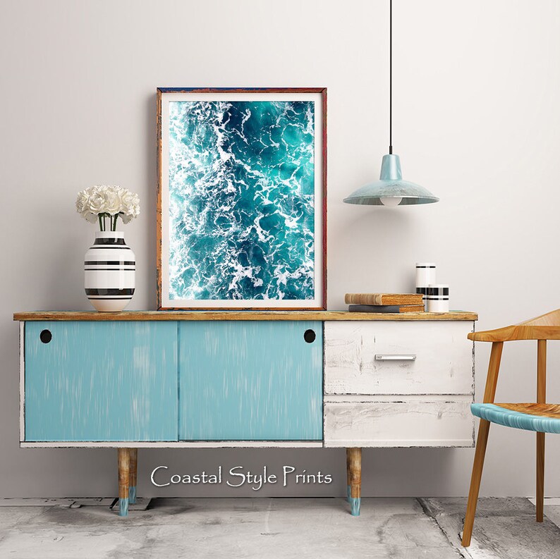 May include: A framed photograph of a blue ocean with white foam waves. The photograph is hanging on a wall above a light blue and white cabinet with a wooden top. The cabinet has two doors and two drawers. There is a white vase with white flowers on the cabinet. The text "Coastal Style Prints" is printed below the cabinet.
