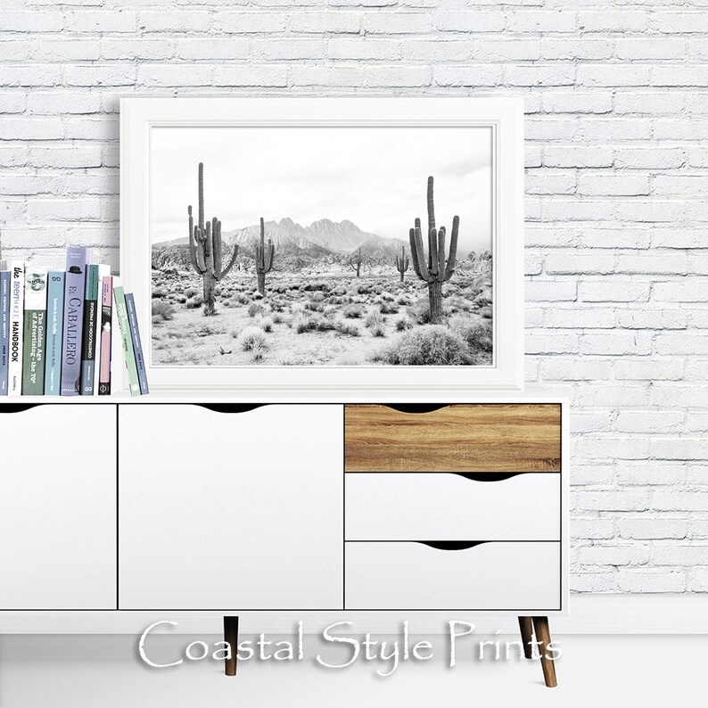 May include: Black and white photograph of a desert landscape with tall cacti in the foreground. The photo is framed and displayed on a white dresser with wooden drawers.