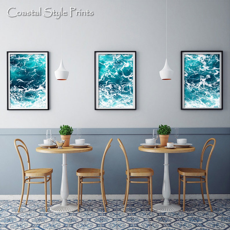 May include: Three framed prints with a vibrant ocean wave design in shades of teal and white hang above a dining table setting. The text "Coastal Style Prints" is in the upper left corner. The room has a blue and white patterned floor.