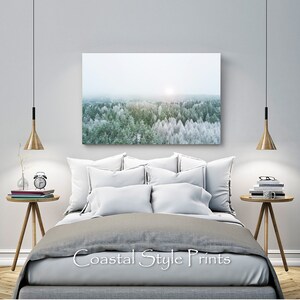 May include: A gray and white bedroom with a large canvas print of a snowy forest scene hanging above a gray bed with white pillows. The bed is styled with a gray duvet and a white throw pillow. There are two wooden side tables with lamps and books on them.