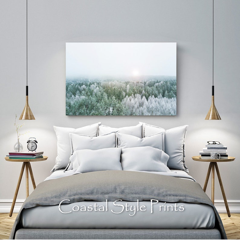 May include: A gray and white bedroom with a large canvas print of a snowy forest scene hanging above a gray bed with white pillows. The bed is styled with a gray duvet and a white throw pillow. There are two wooden side tables with lamps and books on them.