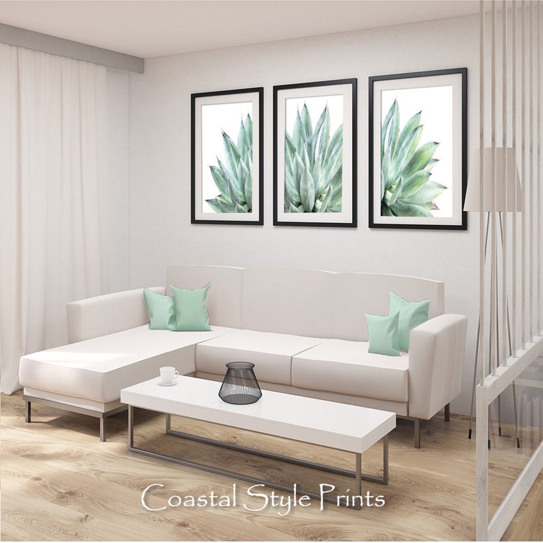 Pu&ograve; includere: Tre stampe incorniciate di una pianta di agave verde su sfondo bianco. Le stampe sono appese a una parete bianca sopra un divano angolare bianco con cuscini di colore verde chiaro. Il divano si trova in un soggiorno con un tavolino da caff&egrave; bianco e un oggetto decorativo in metallo nero sul tavolo.