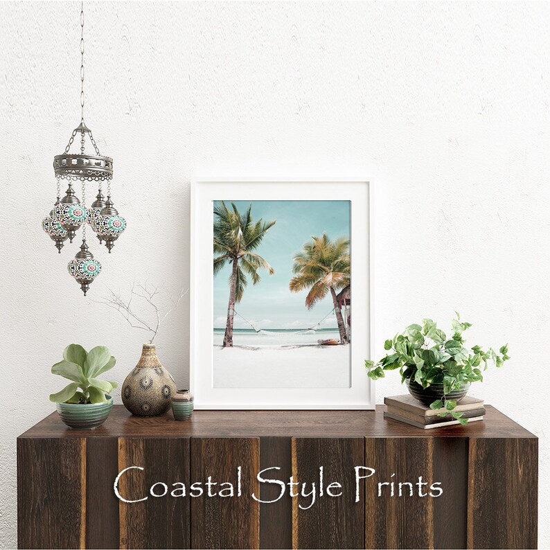 Op de afbeelding: Een witte ingelijste print van een tropisch strandtafereel met twee palmbomen en een hangmat die ertussen hangt. De print staat op een houten plank met een decoratieve lamp, planten en een vaas.