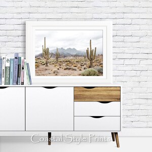 May include: A white framed print of a desert landscape with tall cacti and mountains in the background. The print is hanging on a white brick wall above a white dresser with wooden drawers.