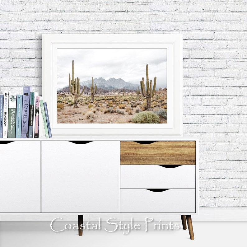 May include: A white framed print of a desert landscape with tall cacti and mountains in the background. The print is hanging on a white brick wall above a white dresser with wooden drawers.