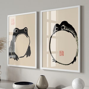 Peut inclure: Deux impressions encadrées avec un fond beige. L'impression de gauche présente un dessin à l'encre noir et blanc d'une grenouille. L'impression de droite présente un dessin à l'encre noir et blanc d'un cercle avec un tampon rouge au centre.