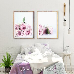 May include: Two framed prints of pink roses on a white background. The prints are hanging on a white wall above a bed with a white and purple patterned comforter.