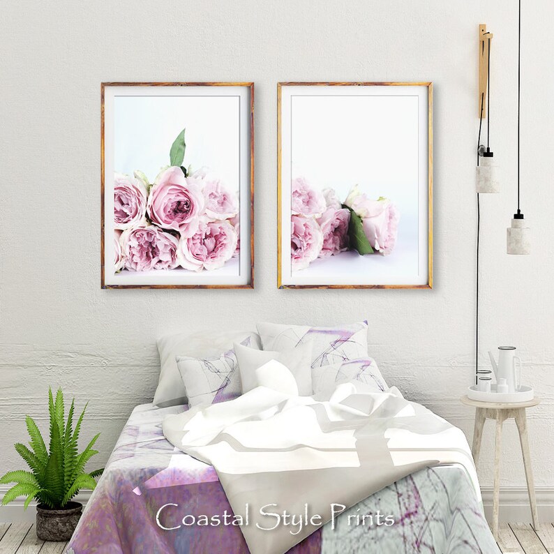 May include: Two framed prints of pink roses on a white background. The prints are hanging on a white wall above a bed with a white and purple patterned comforter.