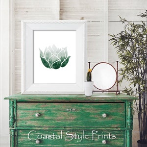 May include: A white frame with a green watercolor print of a lotus flower. The print is on a green dresser with a small cactus plant in a white pot and a round mirror on the dresser.