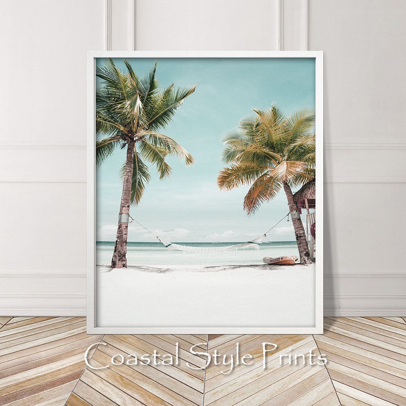 Op de afbeelding: Een wit ingelijste print van een tropisch strand met twee palmbomen en een hangmat die ertussen hangt. Het strand is wit zand en het water is lichtblauw. De lucht is lichtblauw met wat wolken.