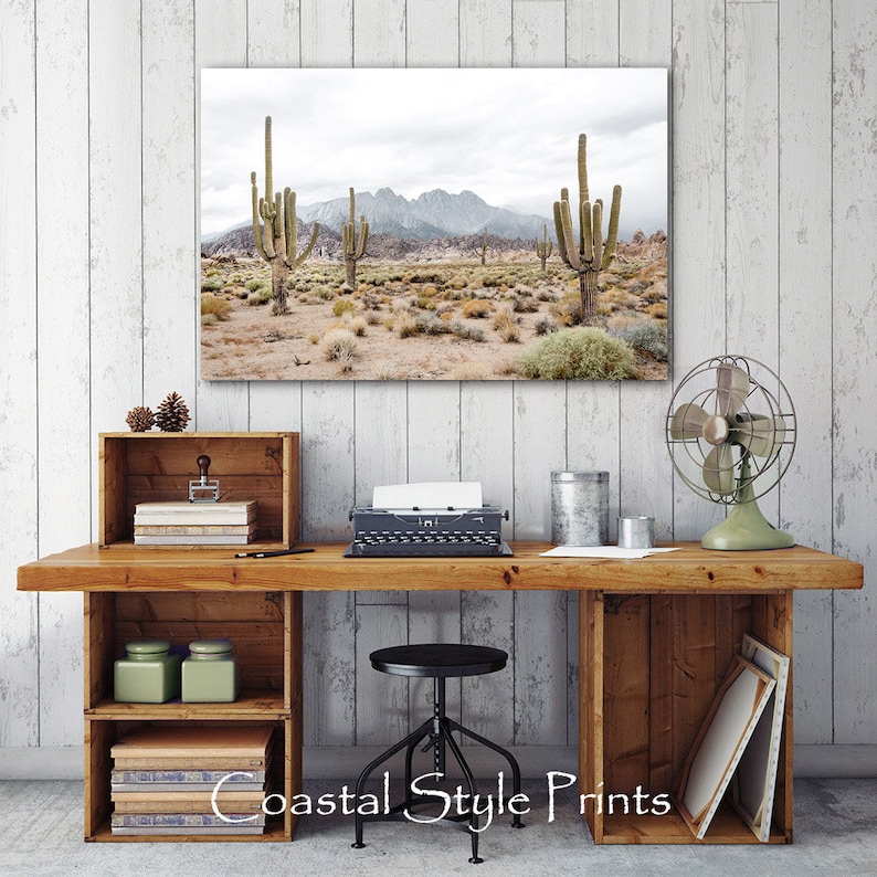 May include: A framed photograph of a desert landscape with tall cacti and a mountain range in the background. The photo is hanging on a white wall above a wooden desk with a typewriter, a fan, and other decorative items.