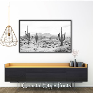 May include: Black and white photograph of a desert landscape with tall cacti and mountains in the background. The photo is framed in a black frame and hanging on a white wall above a black dresser with a gold top.
