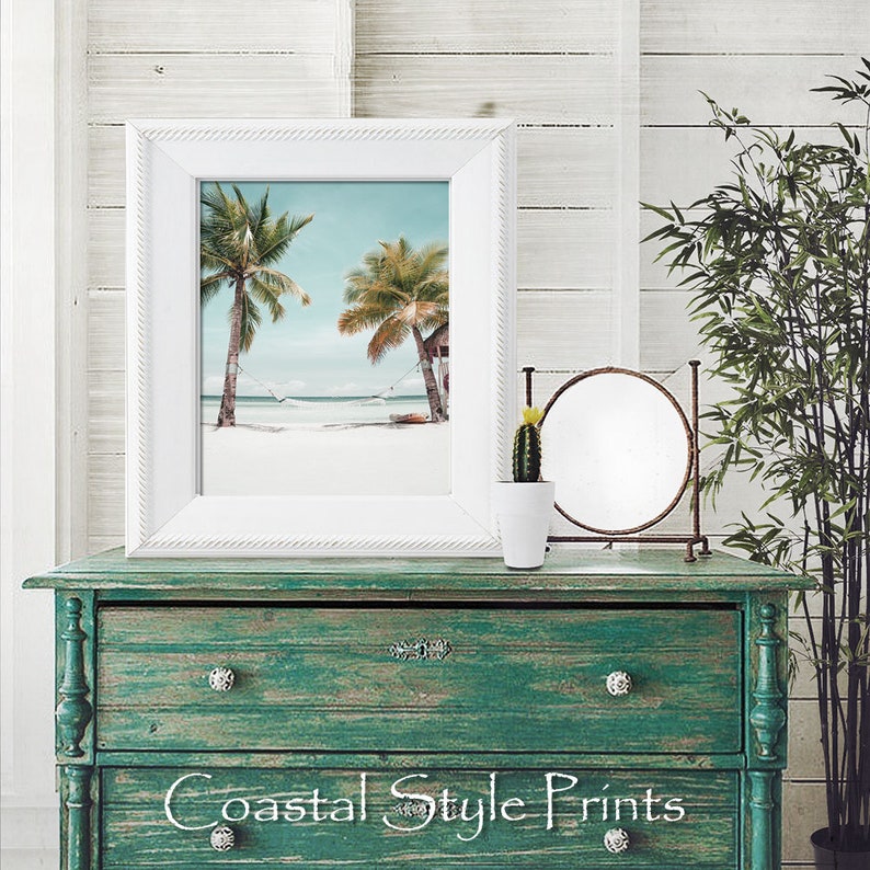Op de afbeelding: Een wit ingelijste print van een tropisch strandtafereel met twee palmbomen en een hangmat die ertussen hangt. De print staat op een groene commode met een ronde spiegel en een kleine cactusplant in een pot.