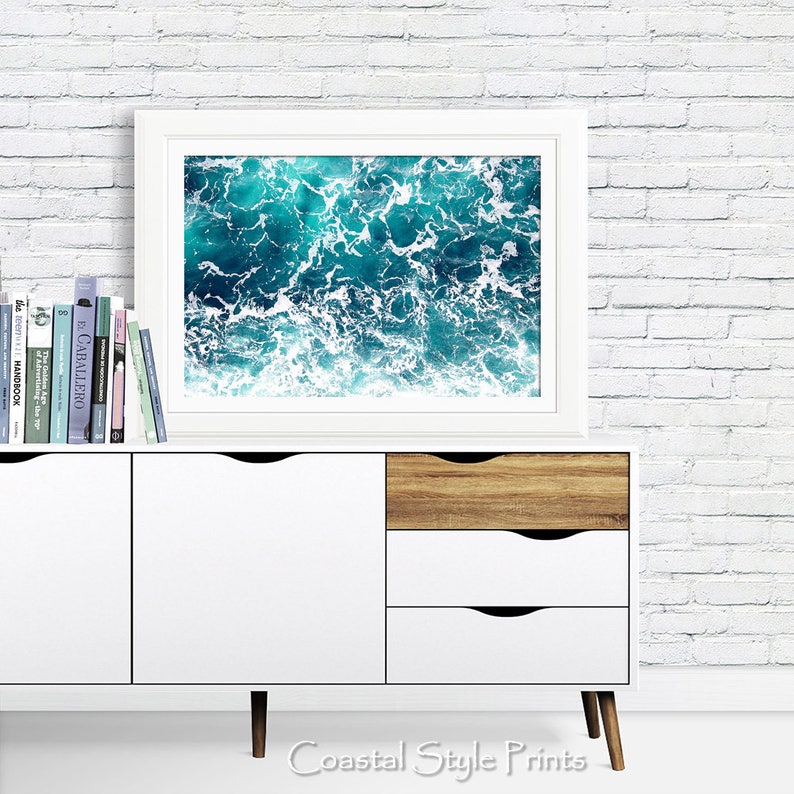 May include: A white framed print of a photograph of blue ocean water with white foam waves. The print is displayed on a white dresser with wooden drawers. The text "Coastal Style Prints" is visible below the dresser.