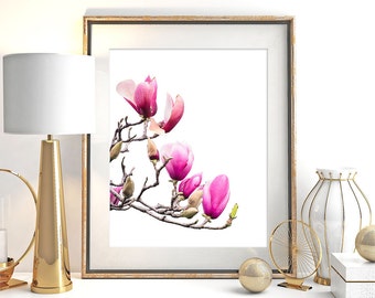 May include: A framed print featuring a close-up of pink magnolia blossoms on a branch. The artwork is displayed on a white surface, alongside a gold lamp, decorative vase, and other gold accents. The frame has a gold finish.