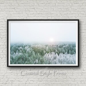 May include: An aerial photograph of a snowy forest with a hazy sky. The trees are a mix of green and white, with the white trees being covered in snow. The sun is shining through the haze, creating a soft glow.