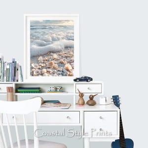 May include: A white desk with a framed photograph of a beach scene with white foam waves and seashells. The desk has a shelf with a toy car and two wooden bird figurines. There is a guitar leaning against the desk.
