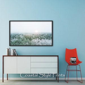 May include: A framed photograph of a snowy forest scene with a sun shining through the trees. The photograph is hanging on a light blue wall above a white cabinet with a wooden top. A red chair with a stack of books is in the foreground.