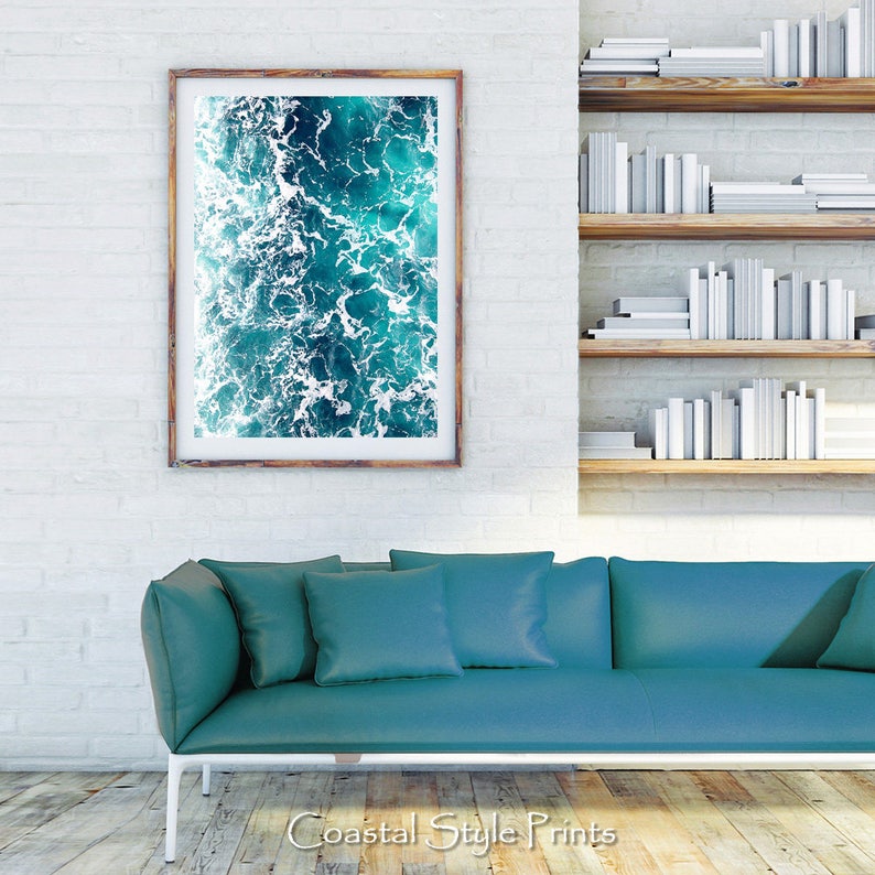 May include: A framed photograph of a turquoise blue ocean with white foam waves. The photograph is hanging on a white brick wall above a teal blue sofa.