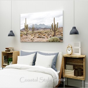 May include: A canvas print featuring a desert landscape with tall cacti and a mountain range in the background. The sky is cloudy and the colors are muted.