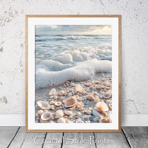 May include: A close-up of white seashells scattered on a sandy beach with foamy ocean waves in the background. The shells are various sizes and shapes, with some partially submerged in the sand.