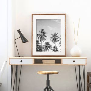 May include: Black and white photograph of palm trees against a cloudy sky in a wooden frame. The framed print is displayed on a wooden desk with a white drawer and black hairpin legs. A black lamp and a white vase with dried branches are also on the desk.