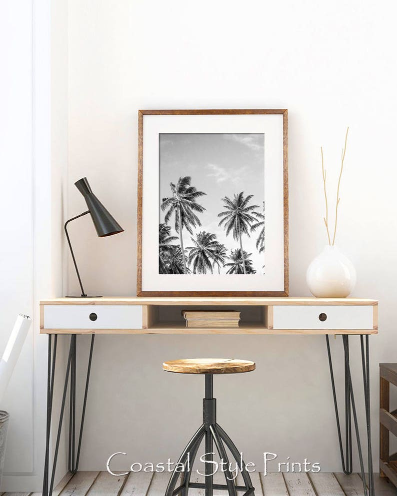 May include: Black and white photograph of palm trees against a cloudy sky in a wooden frame. The framed print is displayed on a wooden desk with a white drawer and black hairpin legs. A black lamp and a white vase with dried branches are also on the desk.
