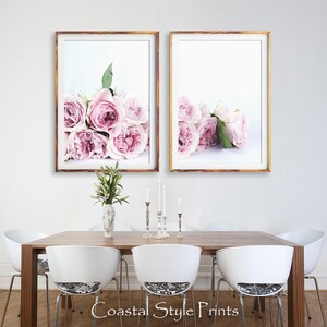 May include: Two framed prints of pink roses on a white background. The prints are hung on a white wall above a wooden dining table with white chairs.