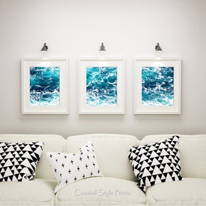 May include: Three framed ocean wave prints in white frames hang above a white sofa with decorative pillows. The prints feature shades of blue and white, depicting the ocean's surface. The pillows have black and white geometric patterns.