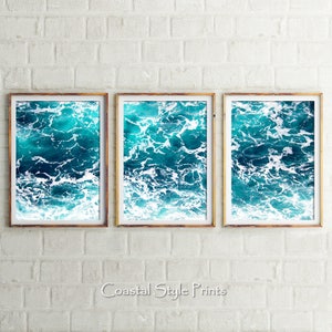 May include: Three framed art prints featuring a close-up view of ocean waves. The water is a vibrant turquoise and deep blue, with white foam. The frames are a warm, light brown color. The text "Coastal Style Prints" is at the bottom.
