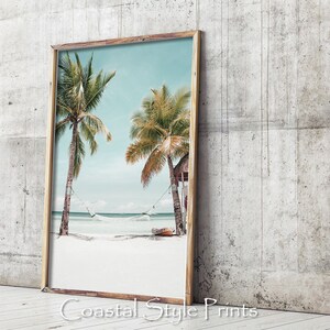 Op de afbeelding: Een ingelijste print van een tropisch strandbeeld met twee palmbomen, een hangmat en een klein hutje. Het strand is wit zand en het water is blauw. De lucht is lichtblauw met witte wolken.