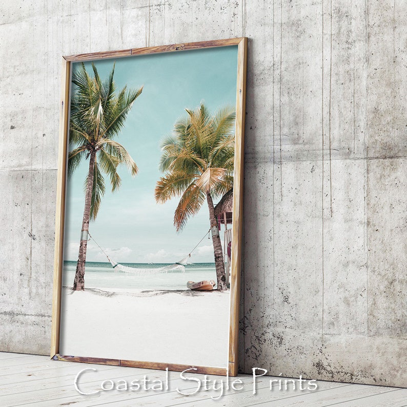 Op de afbeelding: Een ingelijste print van een tropisch strandbeeld met twee palmbomen, een hangmat en een klein hutje. Het strand is wit zand en het water is blauw. De lucht is lichtblauw met witte wolken.