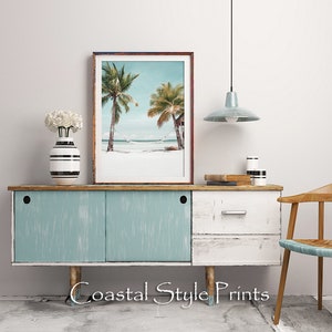 Op de afbeelding: Een ingelijste print van een strandscene met twee palmbomen en een hangmat die ertussen hangt. De print staat op een lichtblauwe en witte commode met een houten blad. De commode heeft twee deuren en een lade. Er staat een witte vaas met witte bloemen op de commode. Een lichtblauwe hanglamp hangt boven de commode.