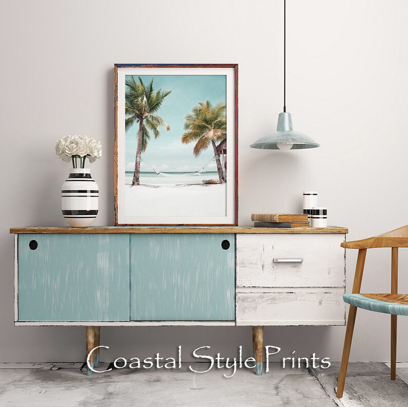 Op de afbeelding: Een ingelijste print van een strandscene met twee palmbomen en een hangmat die ertussen hangt. De print staat op een lichtblauwe en witte commode met een houten blad. De commode heeft twee deuren en een lade. Er staat een witte vaas met witte bloemen op de commode. Een lichtblauwe hanglamp hangt boven de commode.