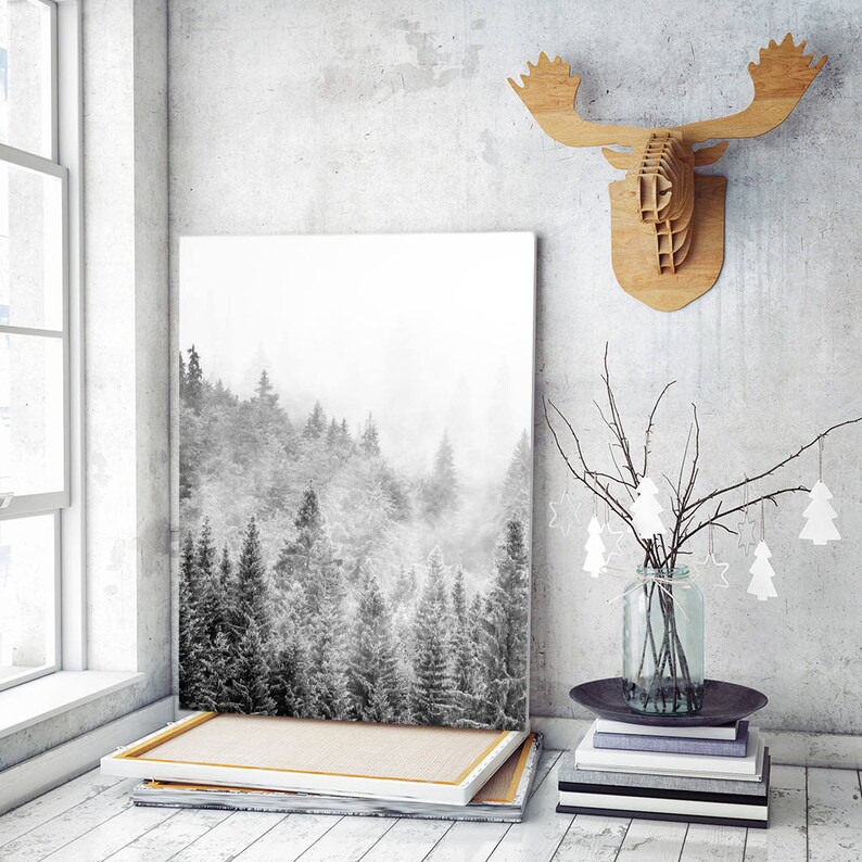 May include: A black and white photograph of a snowy forest scene printed on canvas and framed in a white wood frame. The canvas is hanging on a white wall with a wooden moose head sculpture above it. A vase with a branch of white paper trees is on a stack of books in front of the canvas.