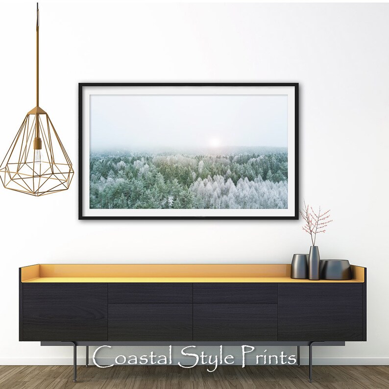May include: A black framed print of a snowy forest scene with a sun shining through the trees. The print is hanging on a white wall above a black dresser with a gold top.