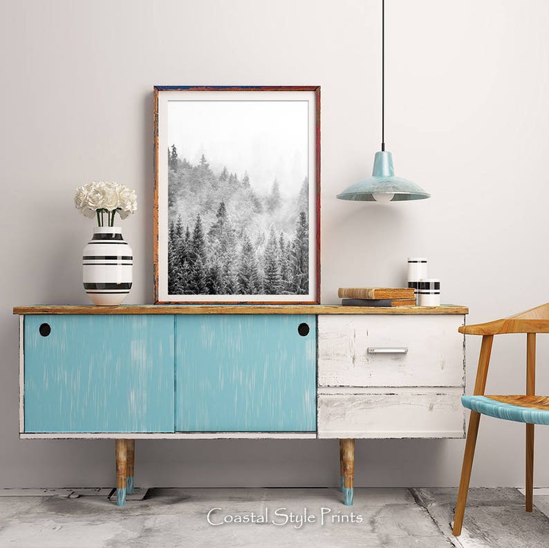May include: A black and white photograph of a foggy forest scene hangs above a light blue and white dresser with a wooden top. The dresser has two drawers and is sitting on four wooden legs. A white vase with black stripes holds a bouquet of white flowers. A light blue pendant lamp hangs from the ceiling above the dresser.