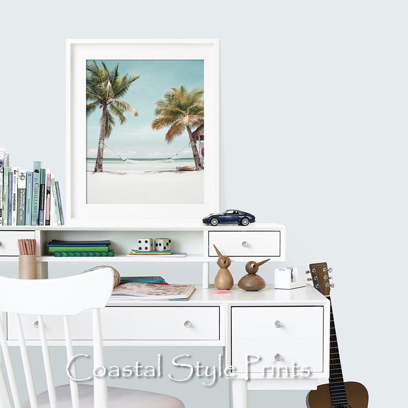 Op de afbeelding: Een witte ingelijste print van een tropisch strandtafereel met twee palmbomen en een hangmat die ertussen hangt. De print hangt aan een witte muur boven een wit bureau met een speelgoedauto, twee houten vogelbeeldjes en een gitaar.