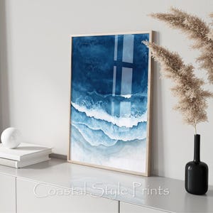 Peut inclure: Impression d'art abstrait représentant une peinture à l'aquarelle bleue et blanche de vagues océaniques. L'impression est encadrée dans un cadre en bois clair.
