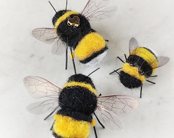 May include: Three needle-felted bumblebee ornaments featuring fuzzy black and yellow bodies, translucent veined wings, and thin black wire legs. One bee is adorned with a small, metallic gold heart-shaped charm on its back.